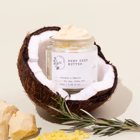 Jar of hemp seed butter inside a coconut with rosemary and more butter on a light background