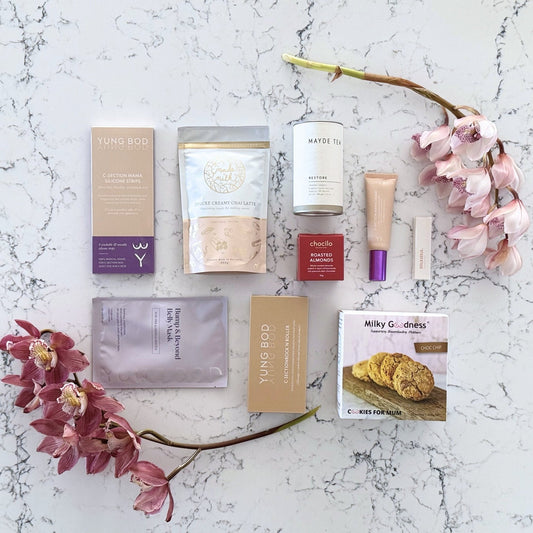 Omoiyari Caesarian Care Luxe gift hamper box - Organic healing tea, C-Section Silicon strips, Scar cream, C-Section skin roller, lactation cookies, chair latte, Essentials oil roller and roasted almonds chocolate with pink orchid flowers on a marble surface