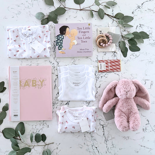 Omoiyari Hello Baby Girl Deluxe Gift hamper bod - Baby gift set with pink plush toy, Marquise baby clothes, teething ring on a marble surface with green leaves.