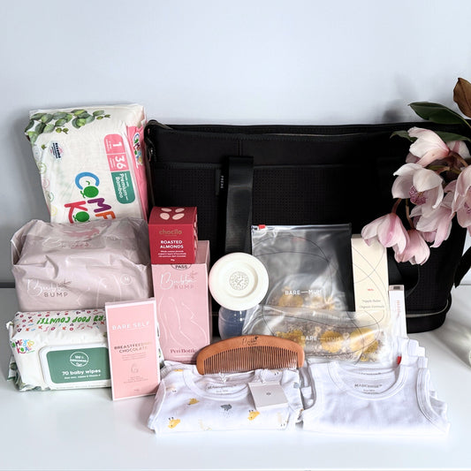 Omoiyari Hospital Bag Luxe with Prene Saturday Bag filled with Marquise babywear, bamboo nappies and wipes, perineal bottle, nipple cream, Essentials Oils roller, Postpartum disposable underwear, bra cooling inserts and chocolate on a marble surface with flowers.