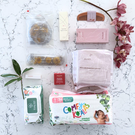 Omoiyari Hospital Bag gift hamper box with bamboo nappies and wipes, perineal bottle, nipple cream, Essentials Oils roller, Postpartum disposable underwear, bra cooling inserts and chocolate on a marble surface with flowers.