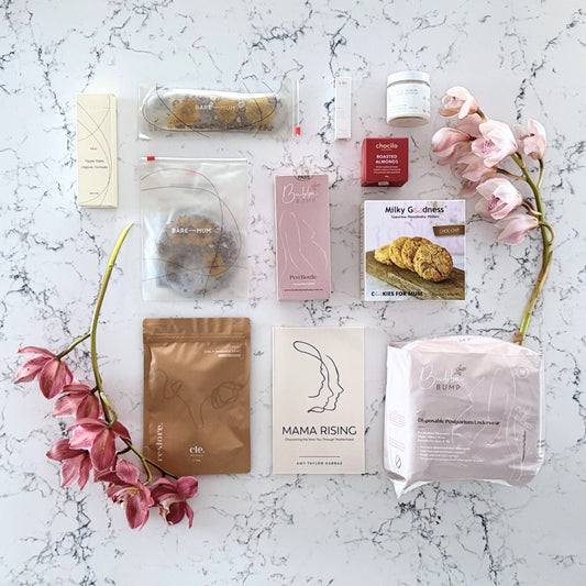 Omoiyari Postpartum Care gift hamper box - Perineal bottle, Sitz bath soak, postpartum underwear, Perineal insert, Lactation cookies, Roasted almond chocolate, nipple balm, Mama Rising book, Essential Oils roller, with pink orchid flowers on a marble surface