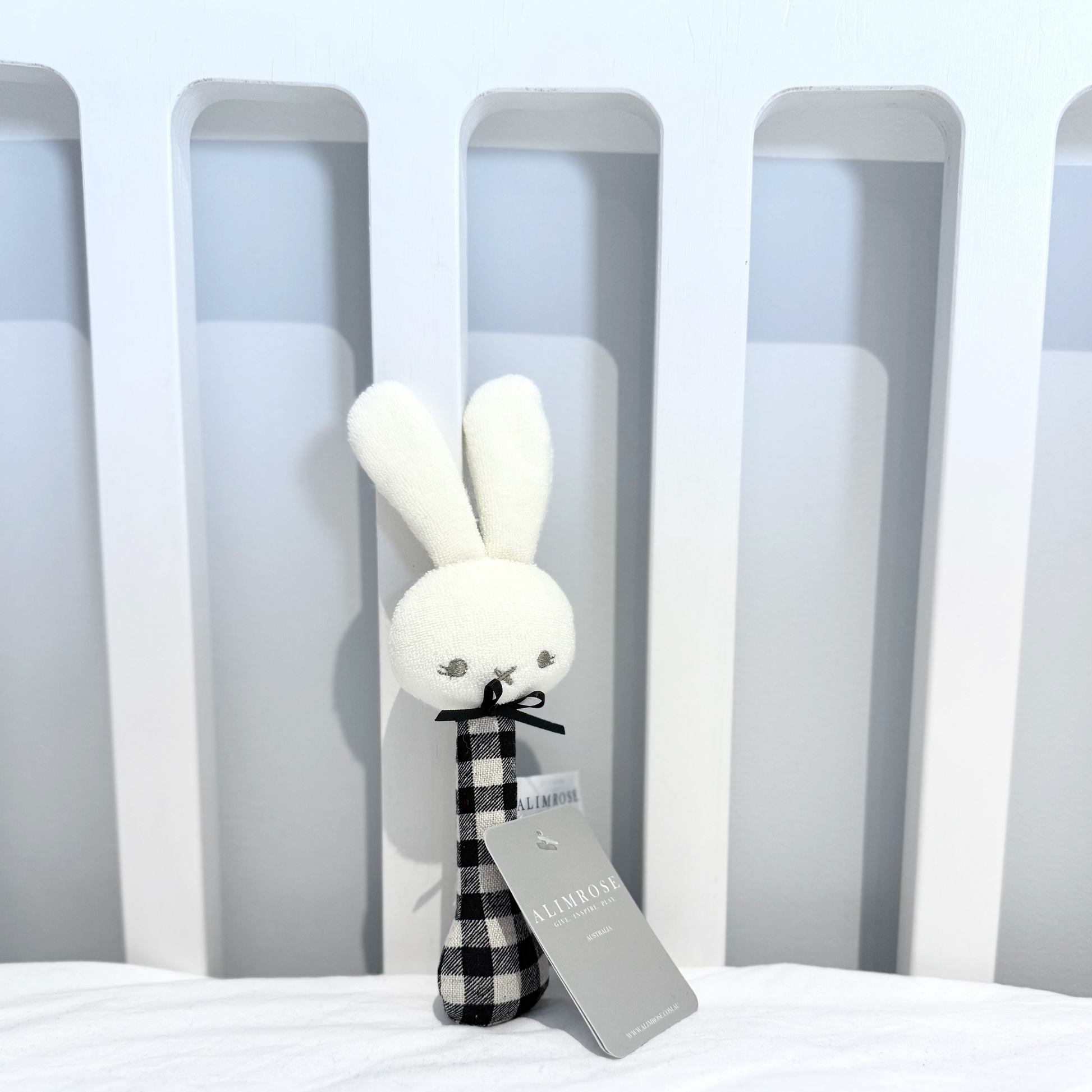 Alimrose White rabbit plush toy with a black and white checkered scarf on a white surface with gray vertical lines in the background.