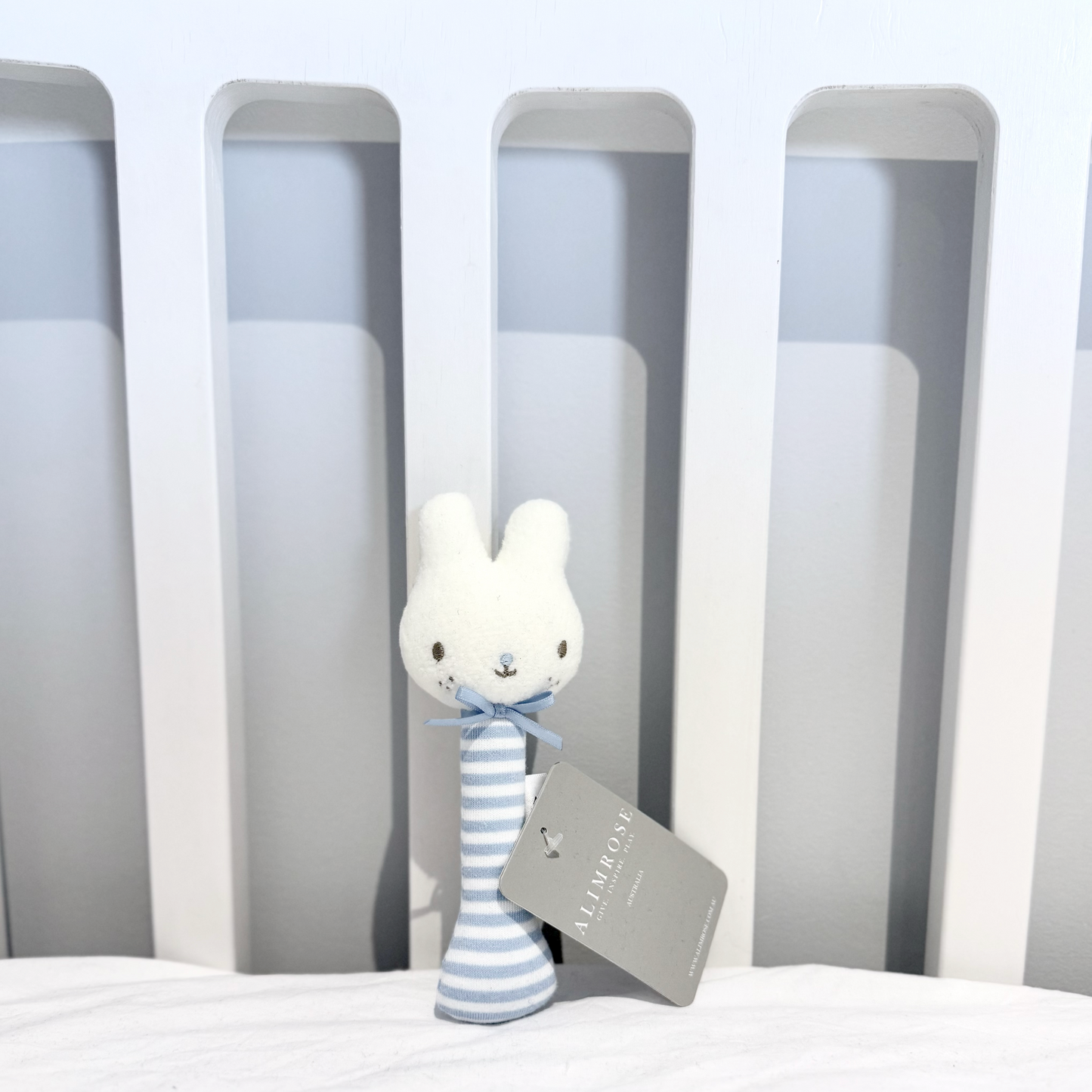 Plush toy with a striped blue body and Alimrose tag on a bed with a neutral headboard