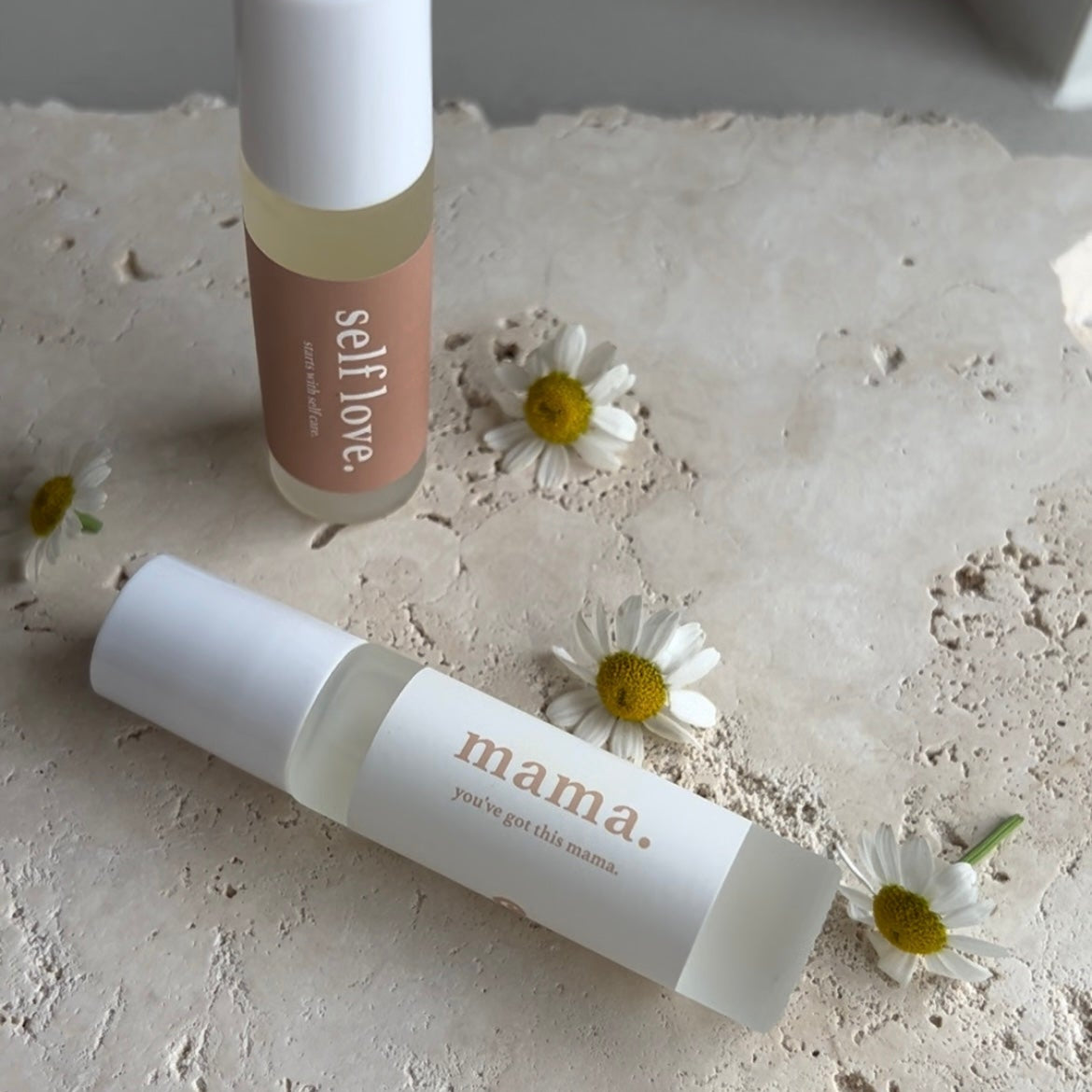 Two skincare products labeled 'self love' and 'mama' on a textured surface with small flowers.