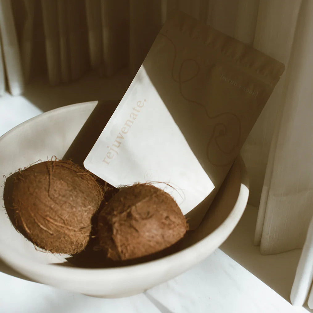 Two coconuts in a white bowl with Cle Naturals Rejuvenate bath soak and a blurred background