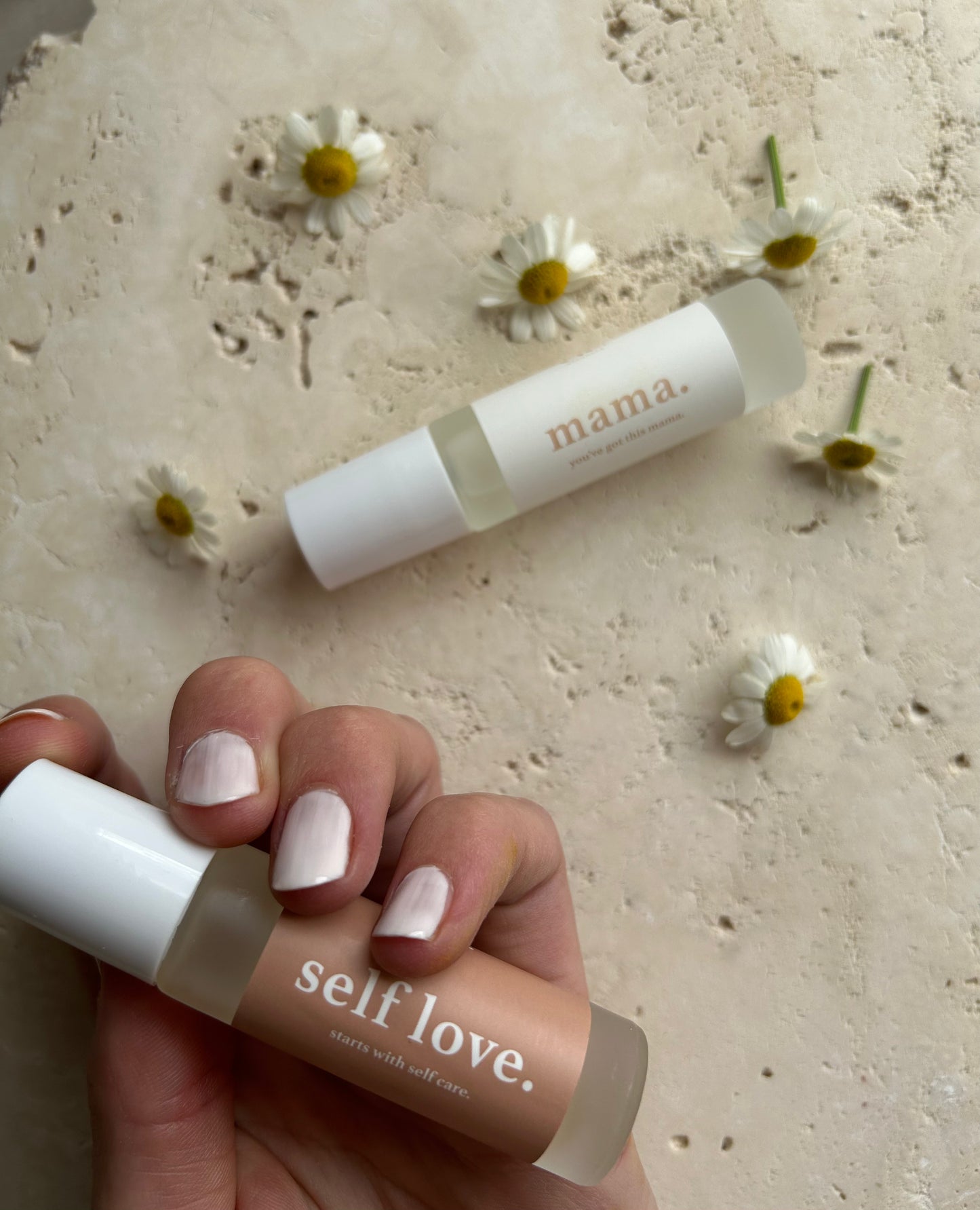 Hand holding a 'self love' product with another one on a textured surface surrounded by small flowers.