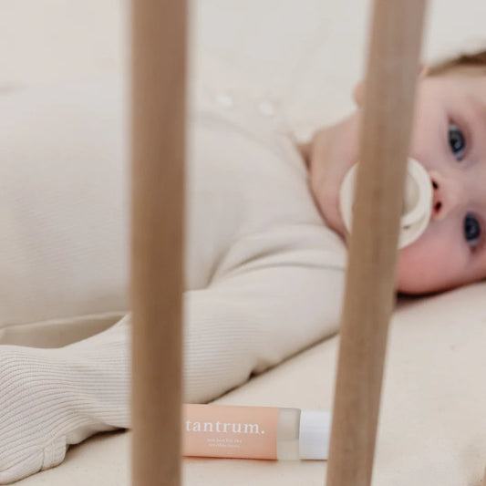 Baby lying in a crib with a pacifier, wearing a white outfit, and a 'tantrum' product visible.