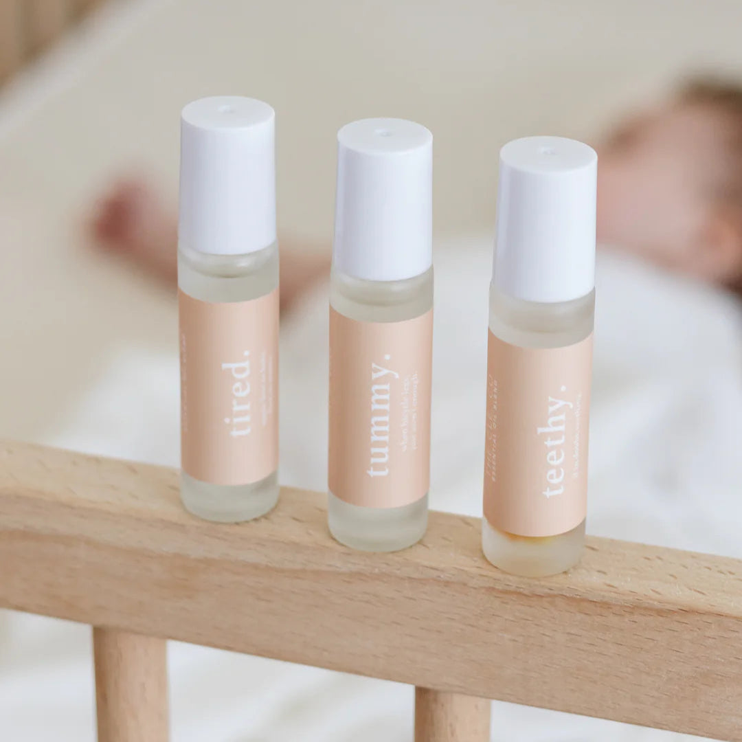 Three Cle Essential Oils Roller bottles labeled 'tired', 'tummy', and 'teethy' on a wooden surface with a blurred background.