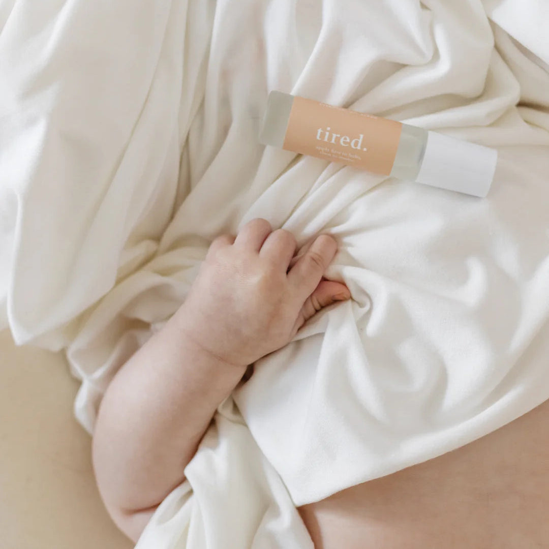 Baby wrapped in a white blanket with Cle Tired Essentials Oil Roller bottle, on a neutral background