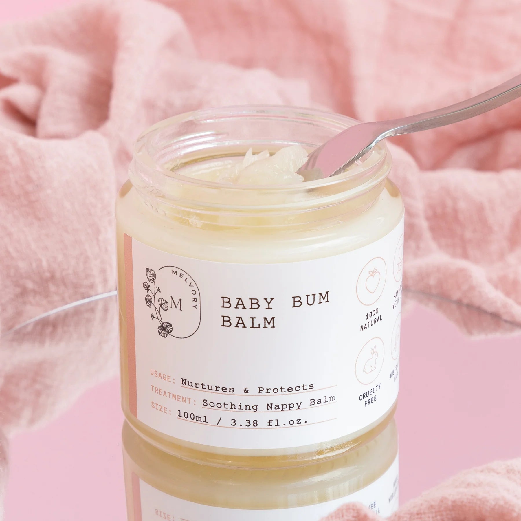 Jar of baby bum balm with a spoon inside on a pink background