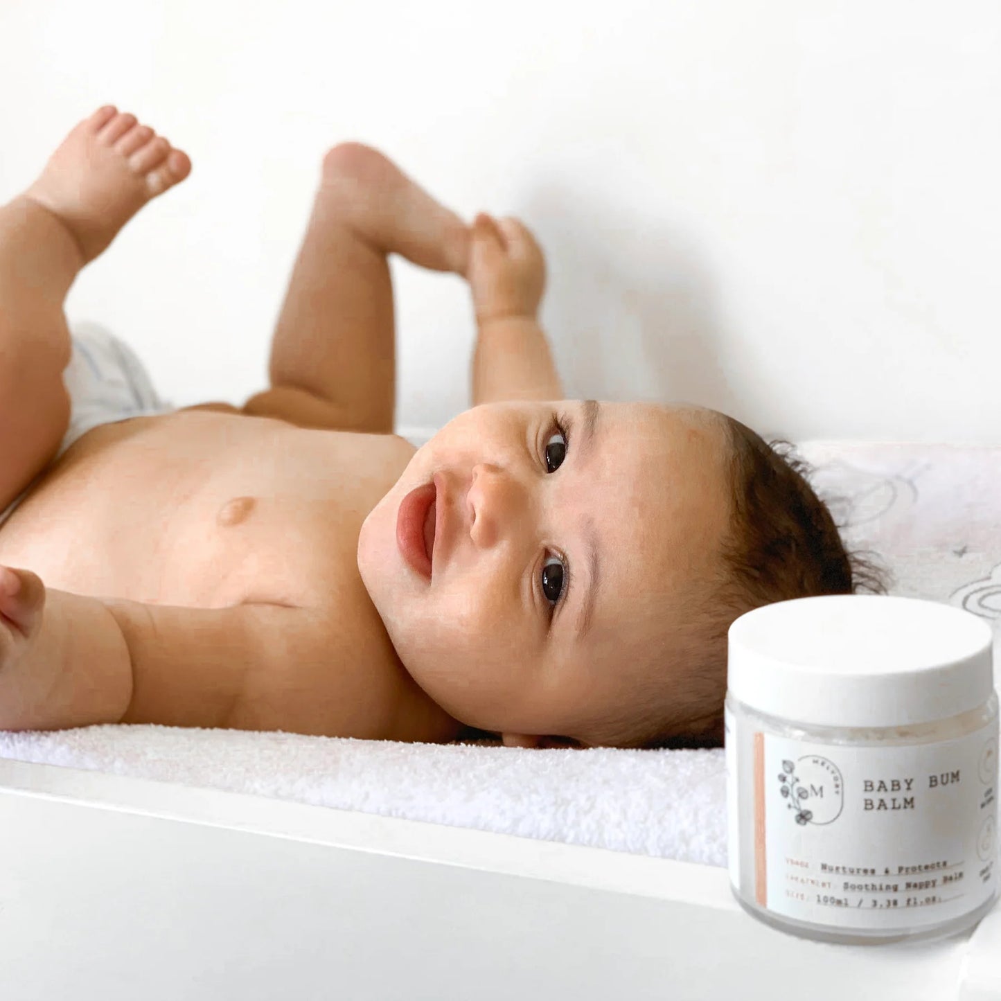 Baby lying on a white blanket with a jar of baby balm next to it.