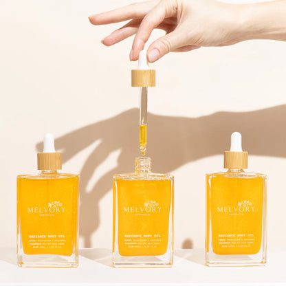 Three bottles of Melvory body oil with a hand holding one bottle's dropper against a neutral background.
