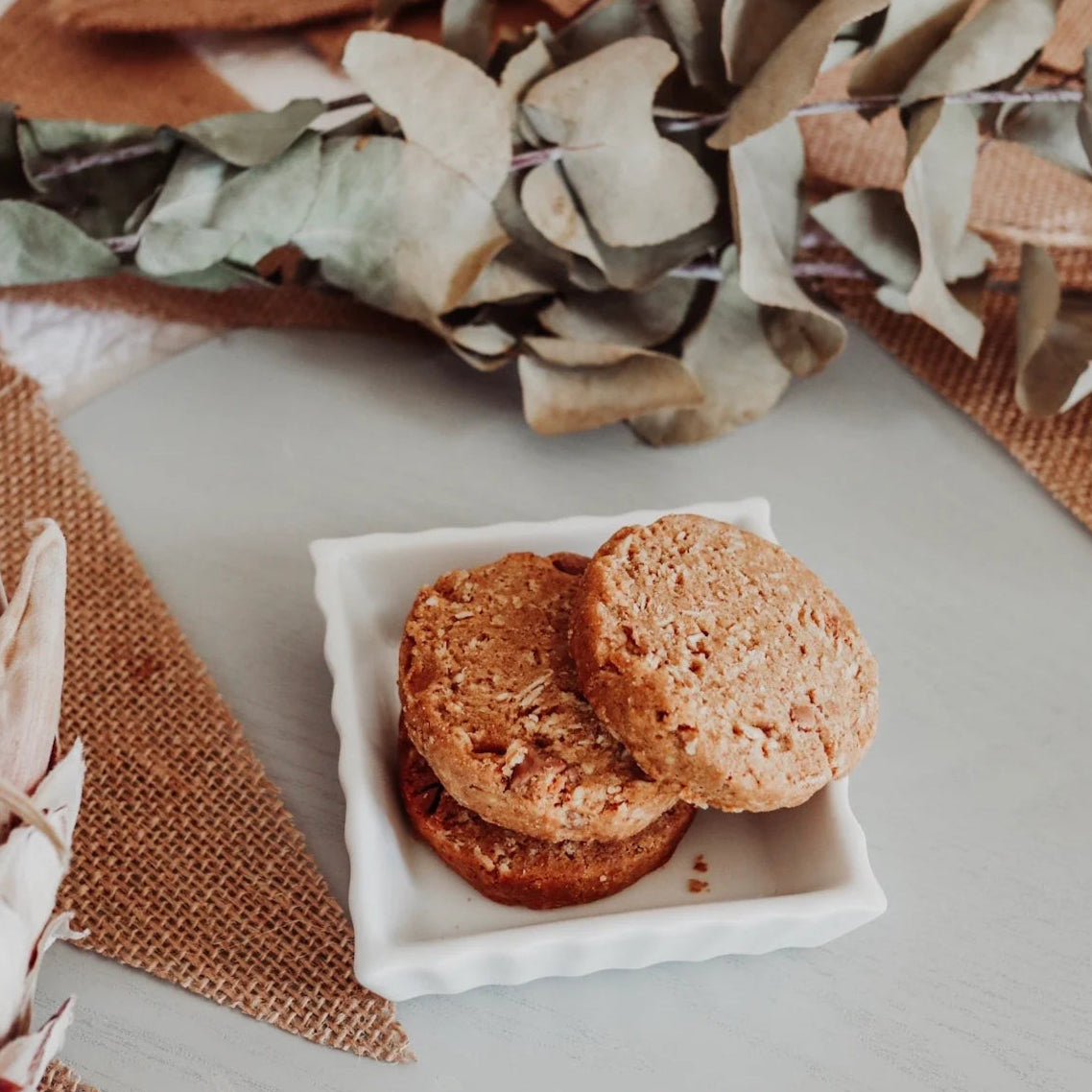 Two cookies on a square white plate with decorative leaves and fabric in the background