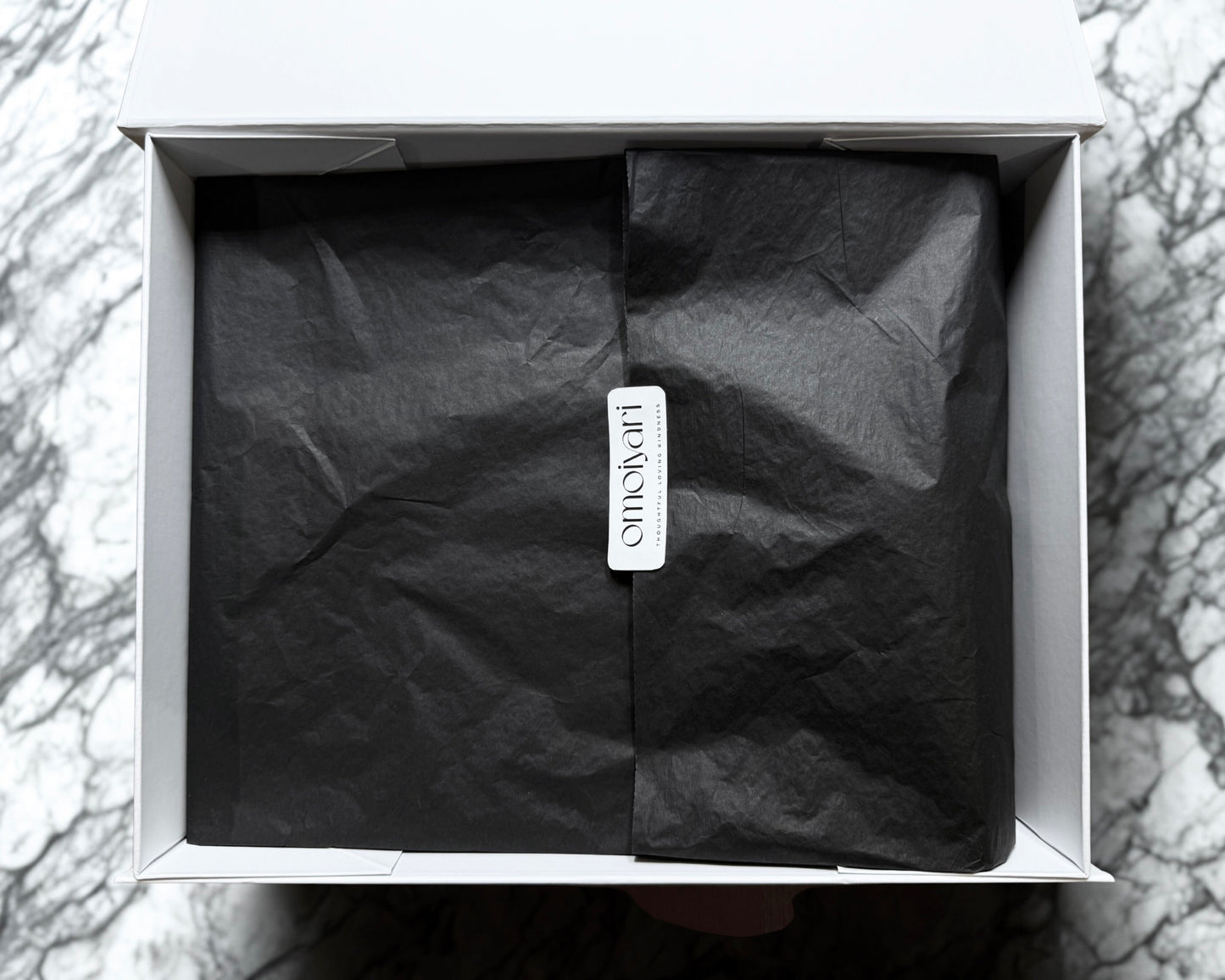 Black tissue paper in a white Omoiyari gift hamper box with a visible Omoiyari brand tag on a marble surface