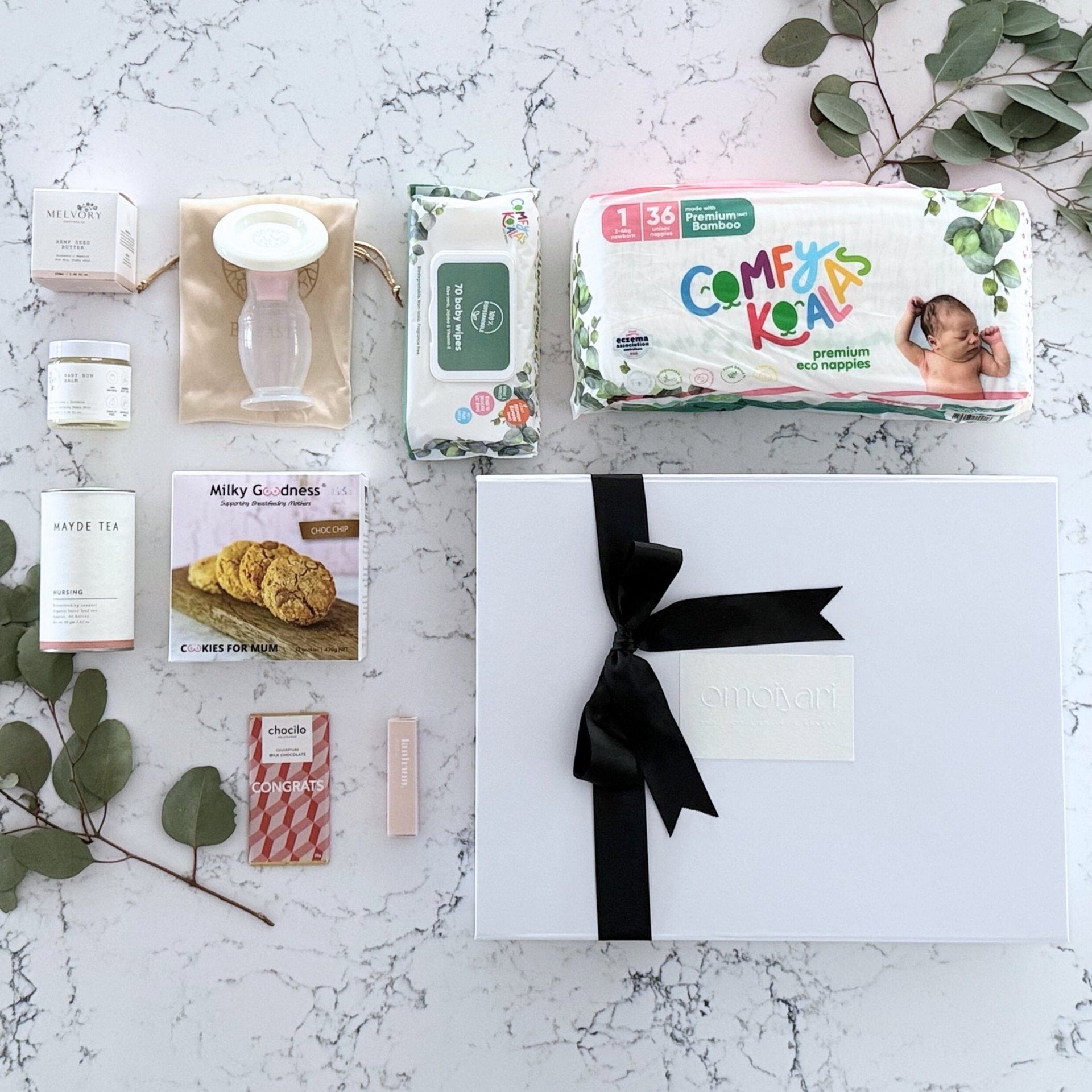 Omoiyari Baby Essentials Luxe Gift hamper box -  nappies, bamboo baby wipes, lactation cookies, chocolate, nursing tea, baby bum balm, essentials oil with green gum leaves on marble table.