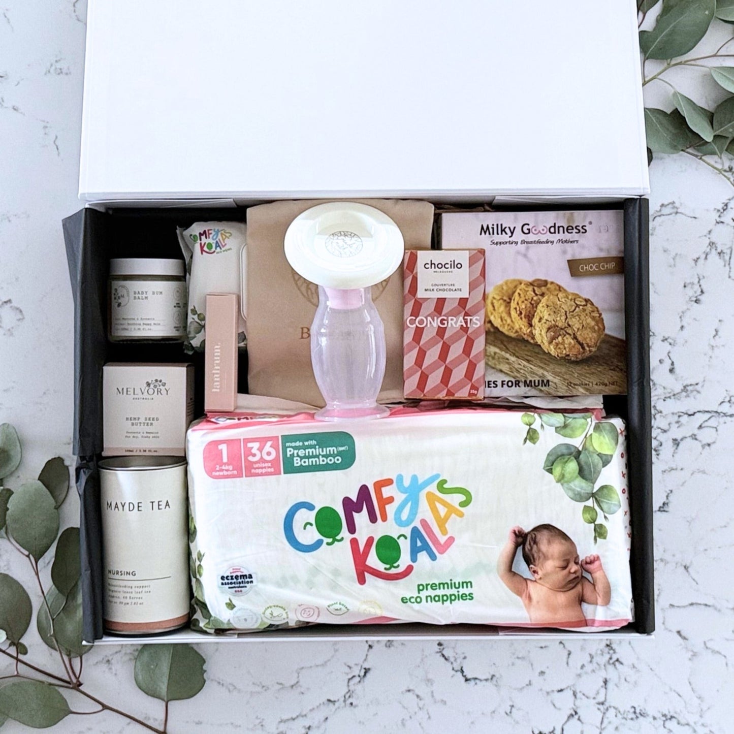 Omoiyari Baby Essentials Luxe Gift hamper box -  nappies, bamboo baby wipes, lactation cookies, chocolate, nursing tea, baby bum balm, essentials oil with green gum leaves on marble table.