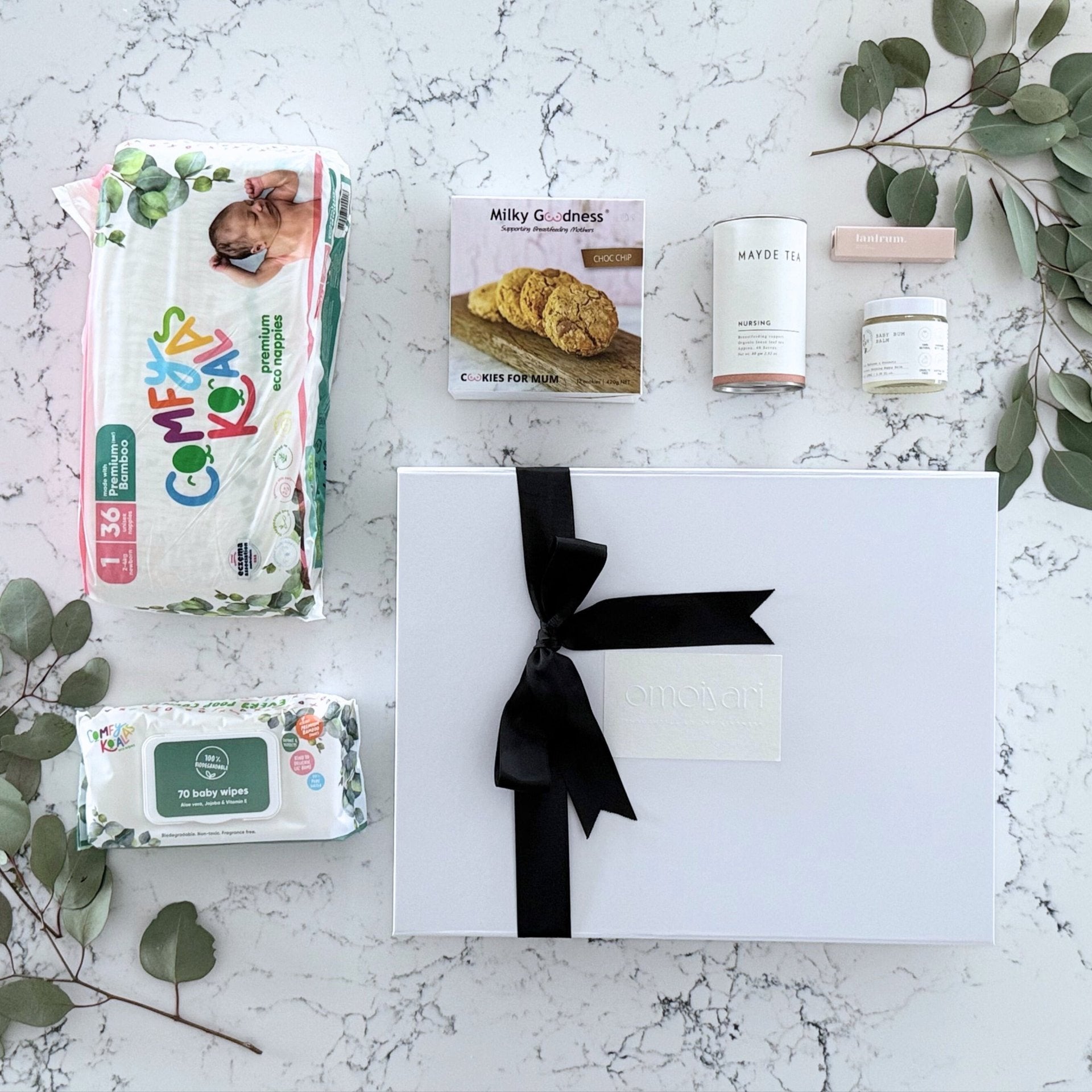 Omoiyari Baby Essentials Gift hamper box -  nappies, bamboo baby wipes, lactation cookies, nursing tea, baby bum balm, essentials oil with green gum leaves on marble table.