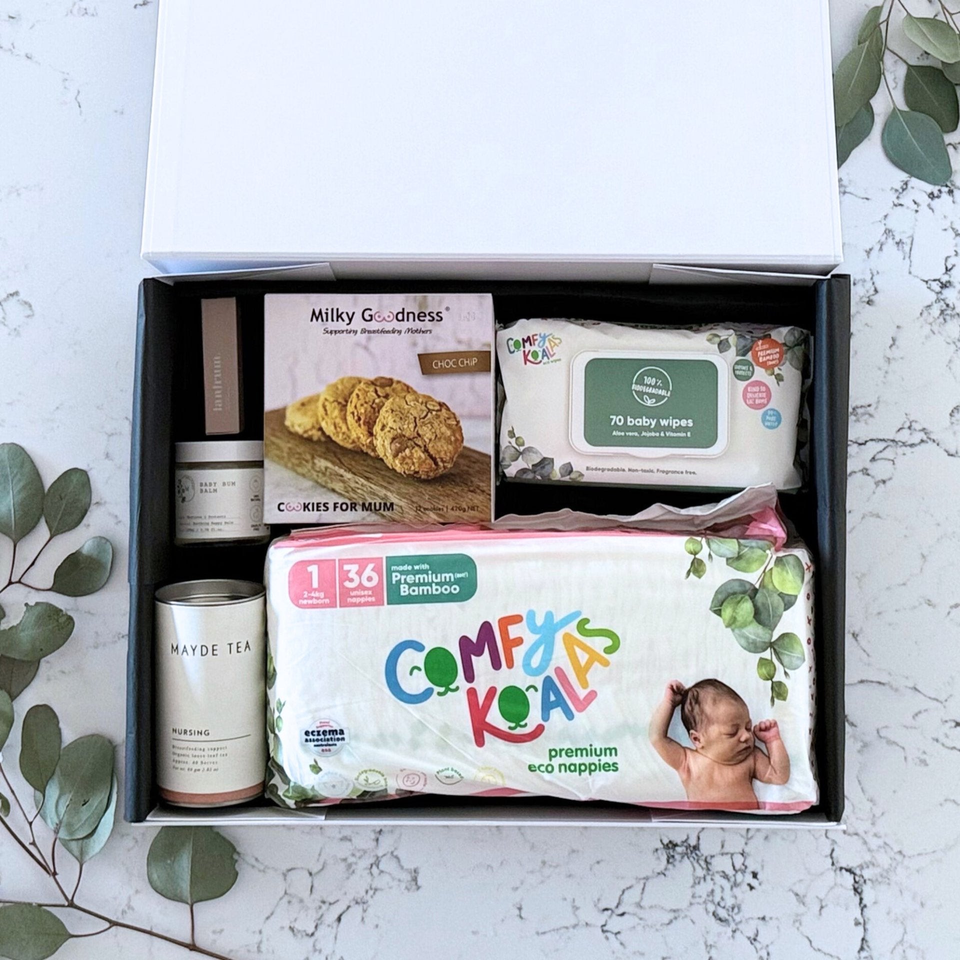 Omoiyari Baby Essentials Gift hamper box -  nappies, bamboo baby wipes, lactation cookies, nursing tea, baby bum balm, essentials oil with green gum leaves on marble table.