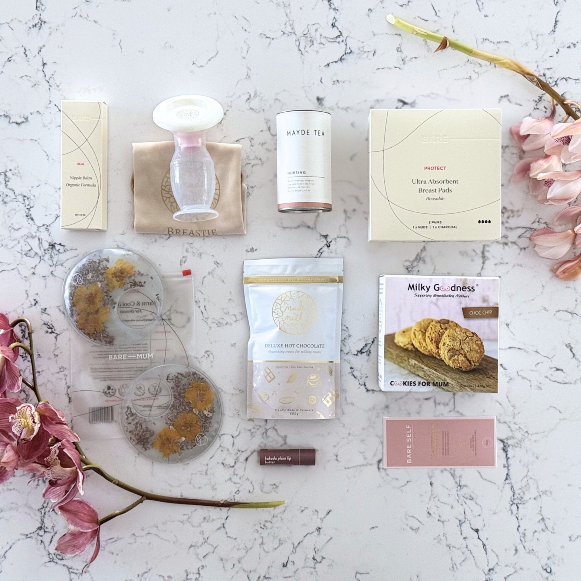 Omoiyari Breastfeeding Essentials Luxe gift hamper box - Lactation cookies, nursing tea, breastfeeding chocolate, Bra cooling inserts, breast pads, lip balm, nipple balm with pink orchid flowers on a marble surface