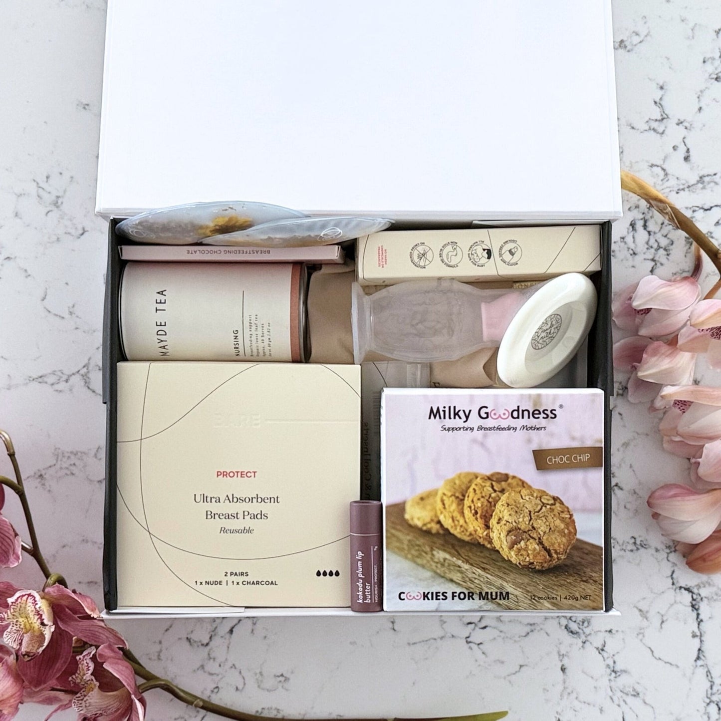 Omoiyari Breastfeeding Essentials Luxe gift hamper box - Lactation cookies, nursing tea, breastfeeding chocolate, Bra cooling inserts, breast pads, lip balm, nipple balm with pink orchid flowers on a marble surface