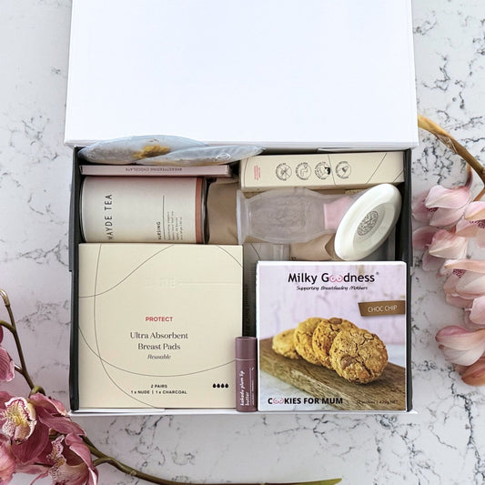 Omoiyari Breastfeeding Essentials Luxe gift hamper box - Lactation cookies, nursing tea, breastfeeding chocolate, Bra cooling inserts, breast pads, lip balm, nipple balm with pink orchid flowers on a marble surface
