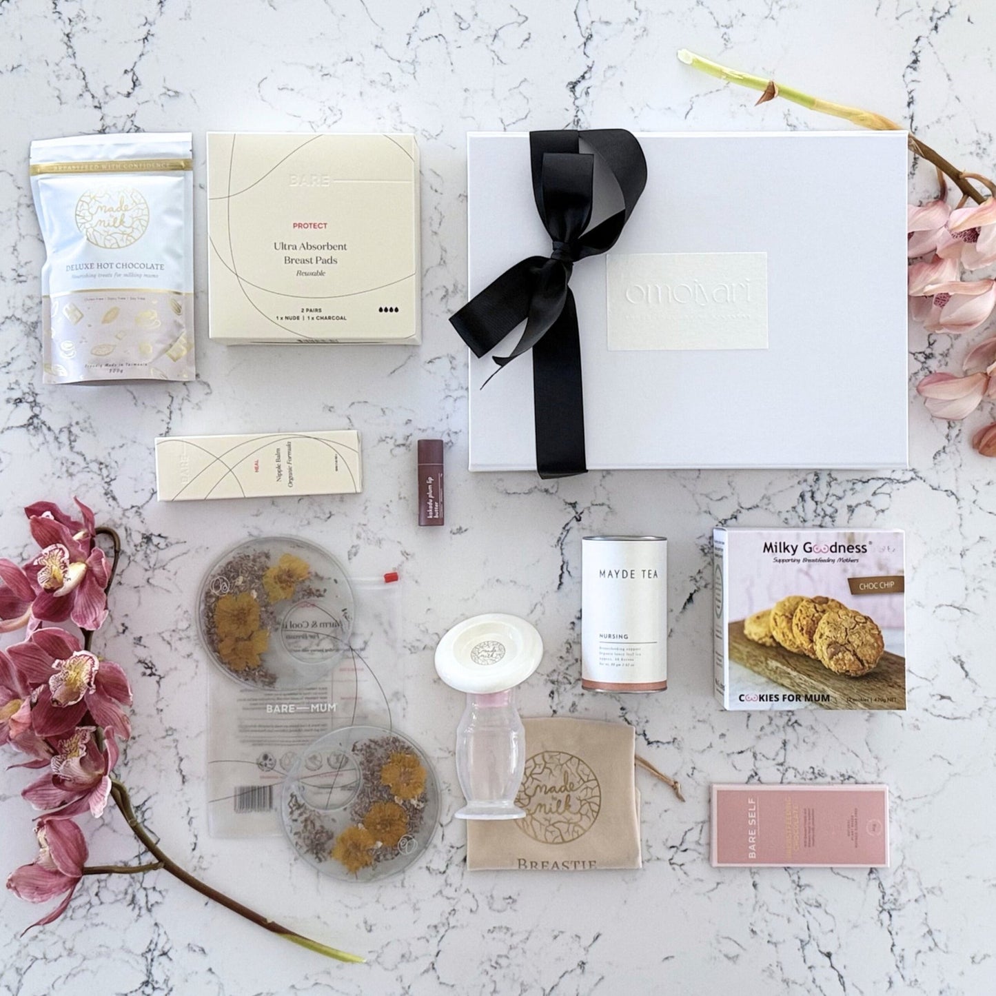 Omoiyari Breastfeeding Essentials Luxe gift hamper box - Lactation cookies, nursing tea, breastfeeding chocolate, Bra cooling inserts, breast pads, lip balm, nipple balm with pink orchid flowers on a marble surface