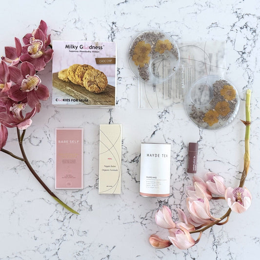 Omoiyari Breastfeeding Essentials gift hamper box - Lactation cookies, nursing tea, breastfeeding chocolate, Bra cooling inserts, lip balm, nipple balm with pink orchid flowers on a marble surface