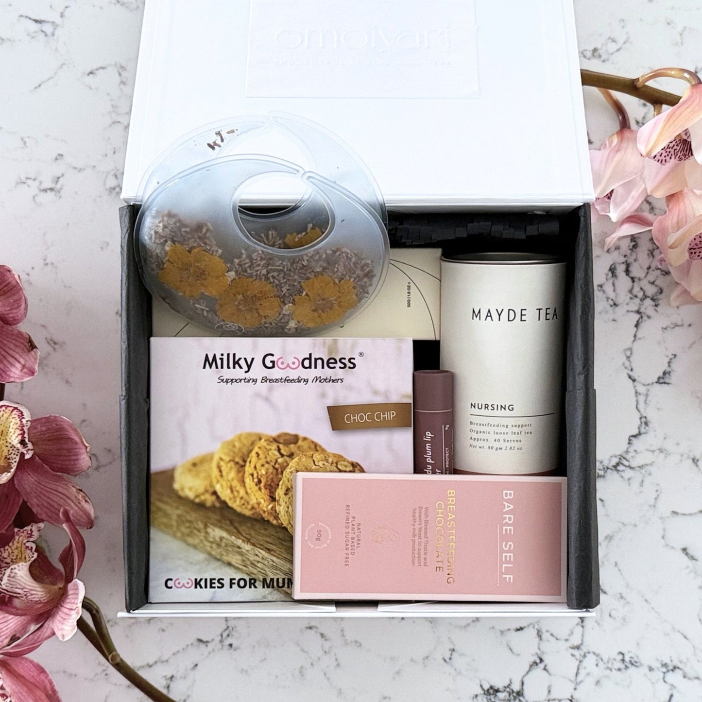 Omoiyari Breastfeeding Essentials gift hamper box - Lactation cookies, nursing tea, breastfeeding chocolate, Bra cooling inserts, lip balm, nipple balm with pink orchid flowers on a marble surface