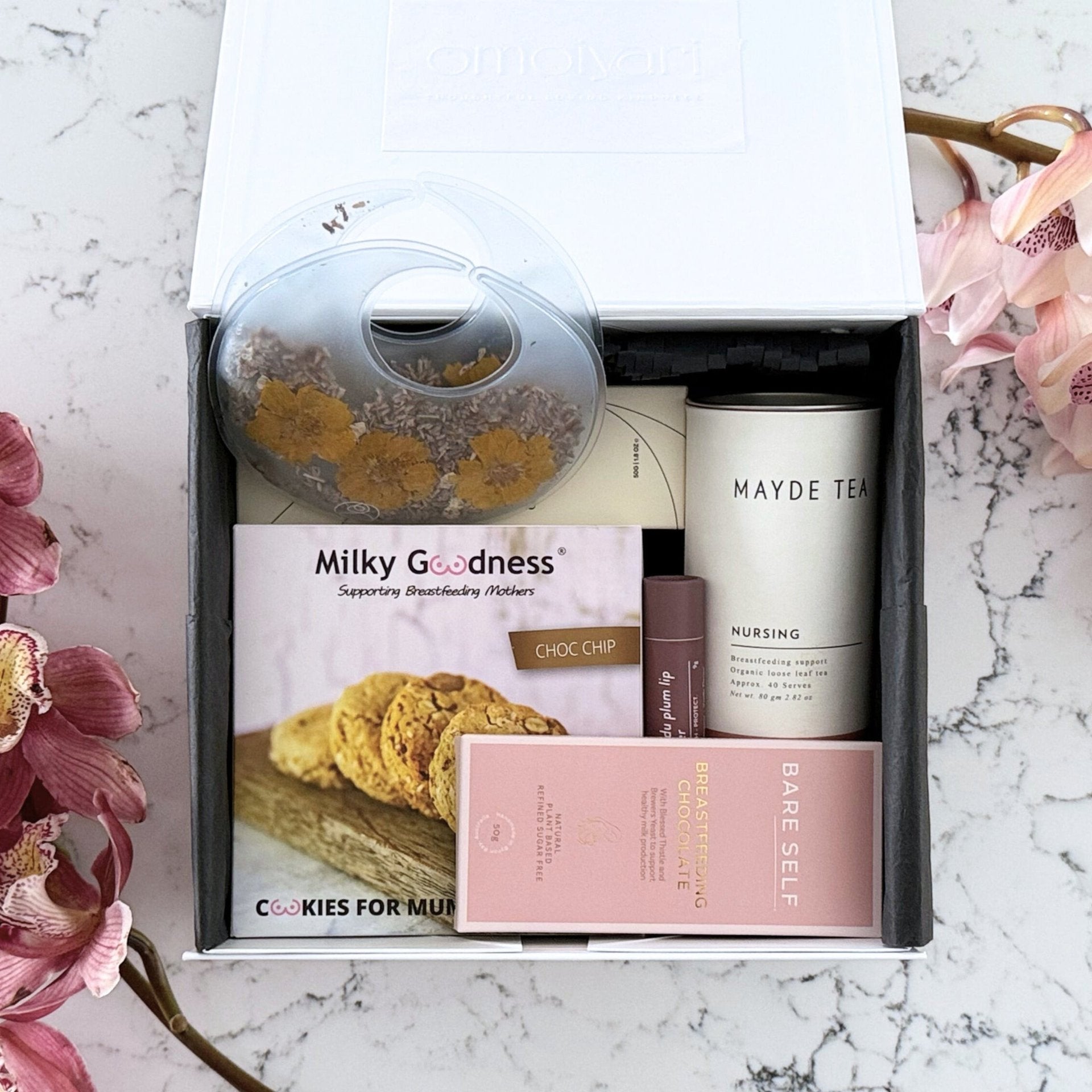 Omoiyari Breastfeeding Essentials gift hamper box - Lactation cookies, nursing tea, breastfeeding chocolate, Bra cooling inserts, lip balm, nipple balm with pink orchid flowers on a marble surface