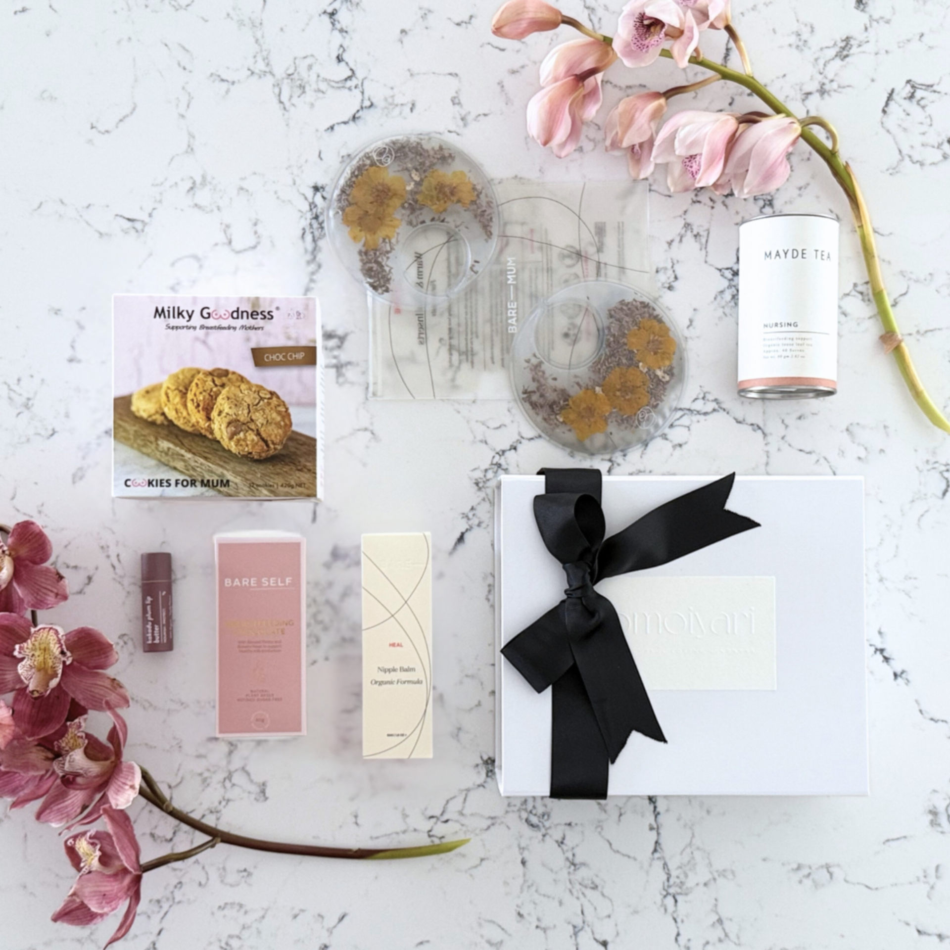 Omoiyari Breastfeeding Essentials gift hamper box - Lactation cookies, nursing tea, breastfeeding chocolate, Bra cooling inserts, lip balm, nipple balm with pink orchid flowers on a marble surface