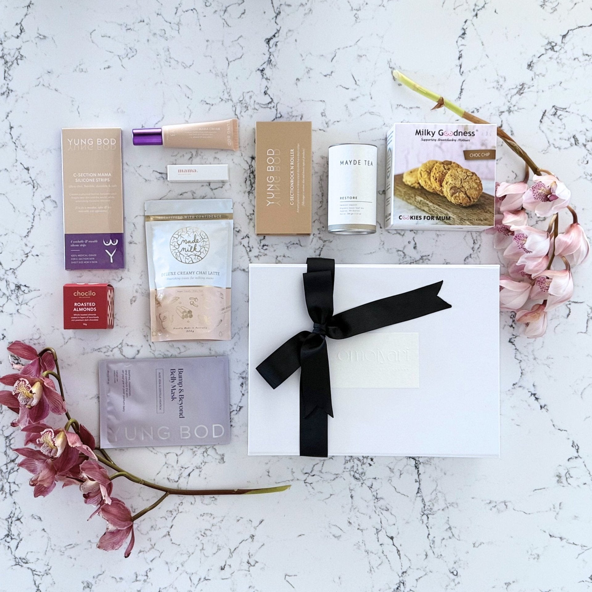 Omoiyari Caesarian Care Luxe gift hamper box - Organic healing tea, C-Section Silicon strips, Scar cream, C-Section skin roller, lactation cookies, chair latte, Essentials oil roller and roasted almonds chocolate with pink orchid flowers on a marble surface
