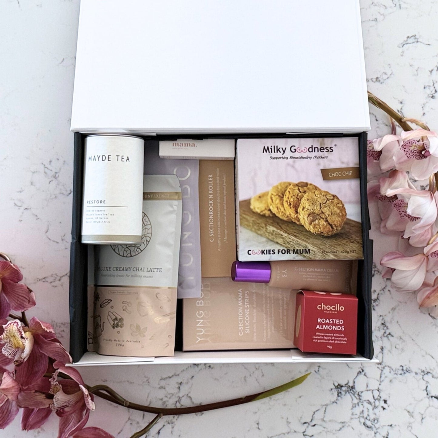 Omoiyari Caesarian Care Luxe gift hamper box - Organic healing tea, C-Section Silicon strips, Scar cream, C-Section skin roller, lactation cookies, chair latte, Essentials oil roller and roasted almonds chocolate with pink orchid flowers on a marble surface