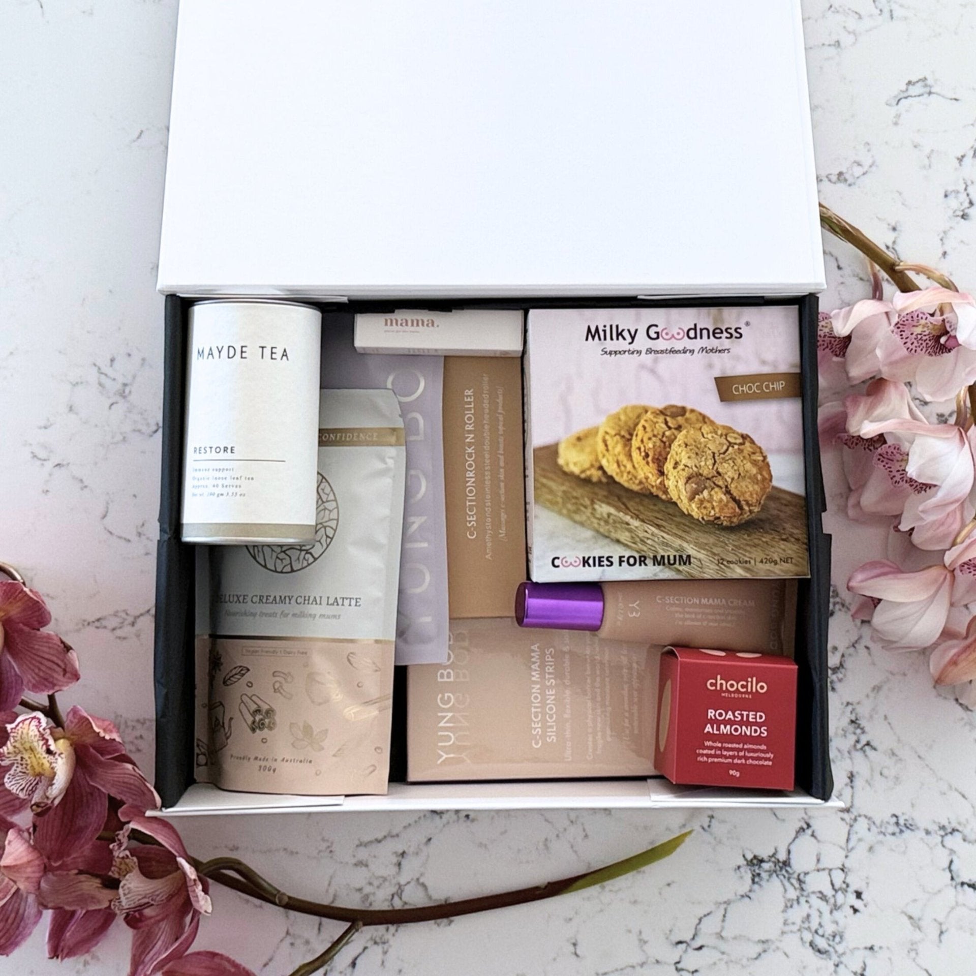 Omoiyari Caesarian Care Luxe gift hamper box - Organic healing tea, C-Section Silicon strips, Scar cream, C-Section skin roller, lactation cookies, chair latte, Essentials oil roller and roasted almonds chocolate with pink orchid flowers on a marble surface