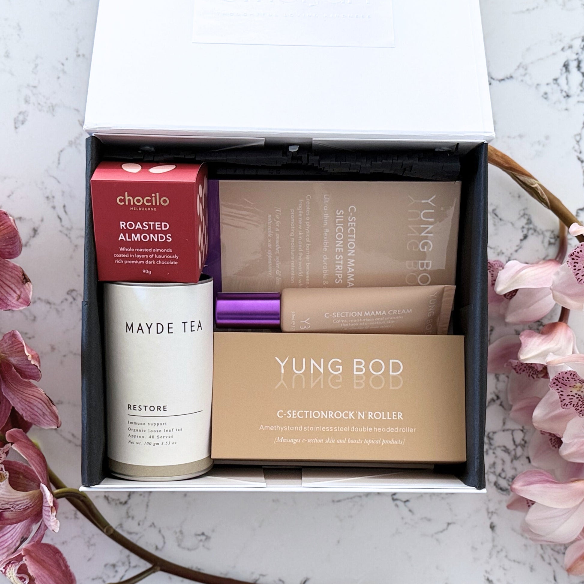 Omoiyari Caesarian Care gift hamper box - Organic healing tea, C-Section Silicon strips, Scar cream, C-Section skin roller and roasted almonds chocolate with pink orchid flowers on a marble surface
