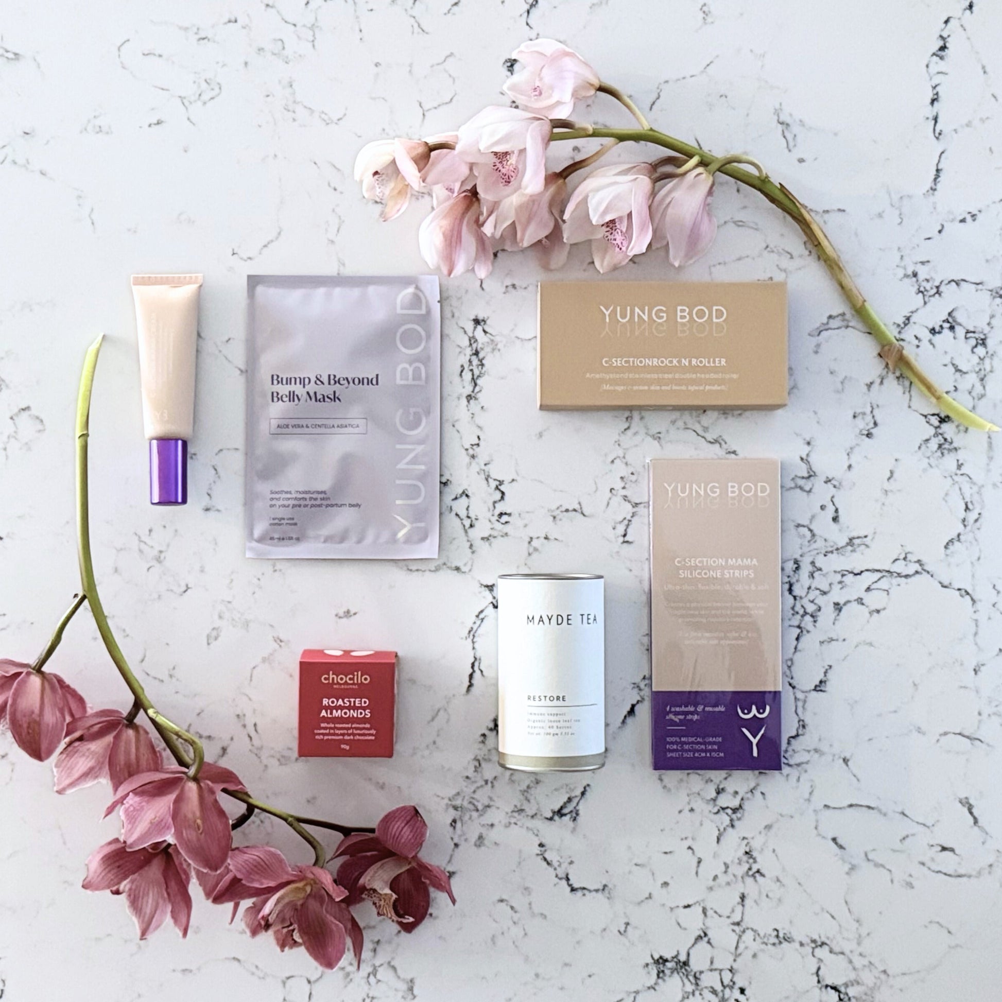 Omoiyari Caesarian Care gift hamper box - Organic healing tea, C-Section Silicon strips, Scar cream, C-Section skin roller and roasted almonds chocolate with pink orchid flowers on a marble surface