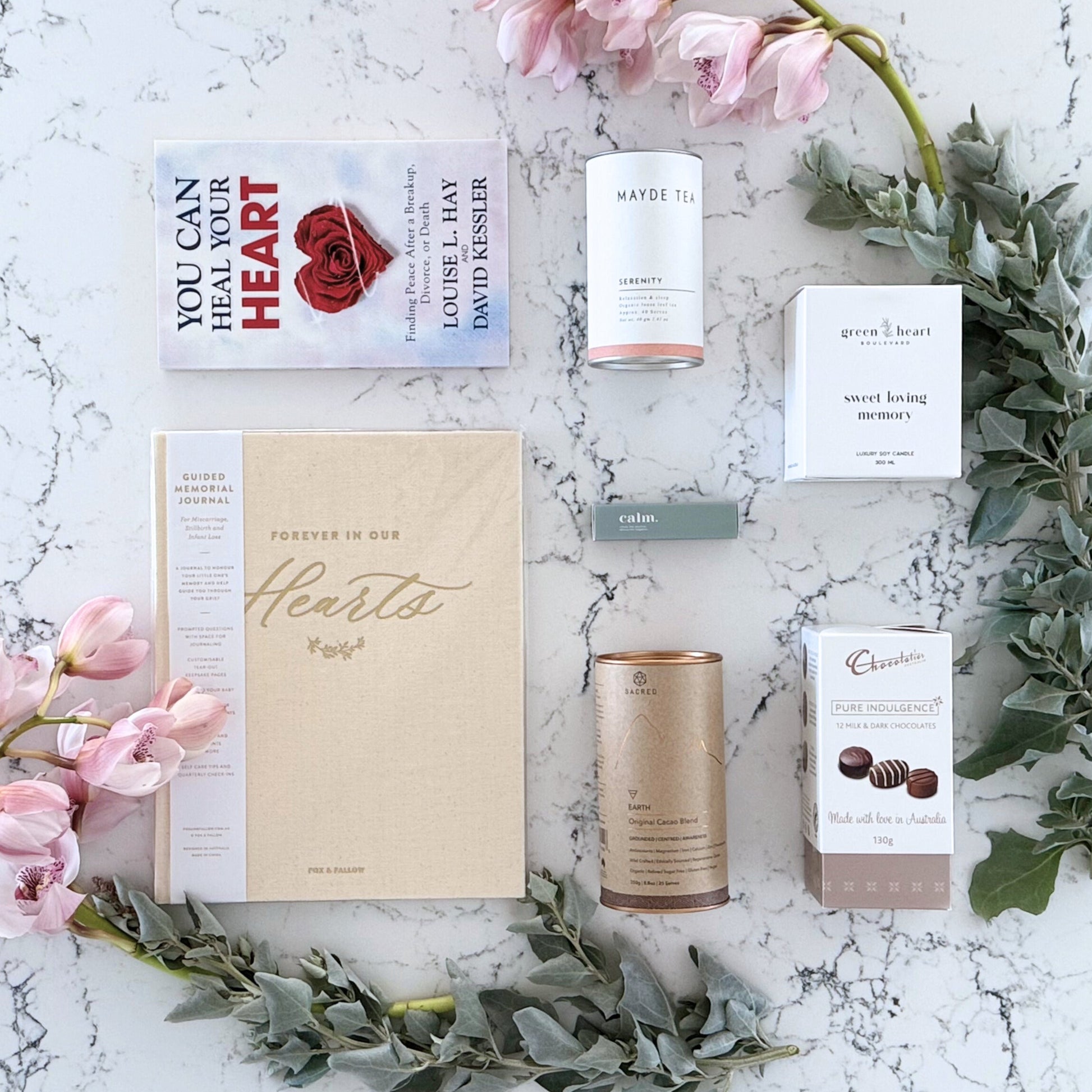 Omoiyari Forever Loved gift hamper box  with self-help book, In memory journal, organic tea, Essentials Oil, Chocolate, and soy candle on a marble surface with flowers.