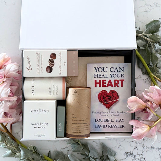 Omoiyari Forever Loved gift hamper box  with self-help book, In memory journal, organic tea, Essentials Oil, Chocolate, and soy candle on a marble surface with flowers.