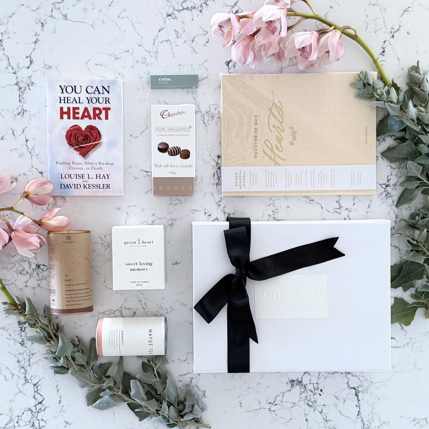 Omoiyari Forever Loved gift hamper box  with self-help book, In memory journal, organic tea, Essentials Oil, Chocolate, and soy candle on a marble surface with flowers.
