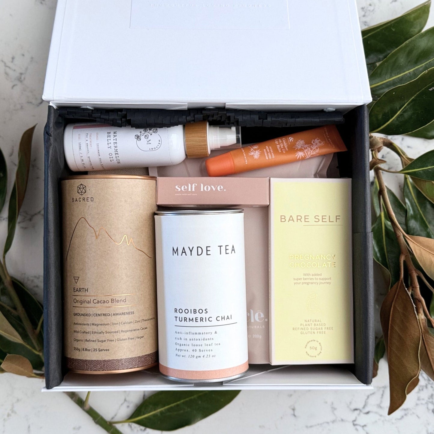 Omoiyari Glow & Grow Luxe gift hamper box - Skincare products, organic cacao and tea, mama pregnancy journal, pregnancy chocolate, stretch mark oil, lip balm and green magnolia leaves on a marble surface