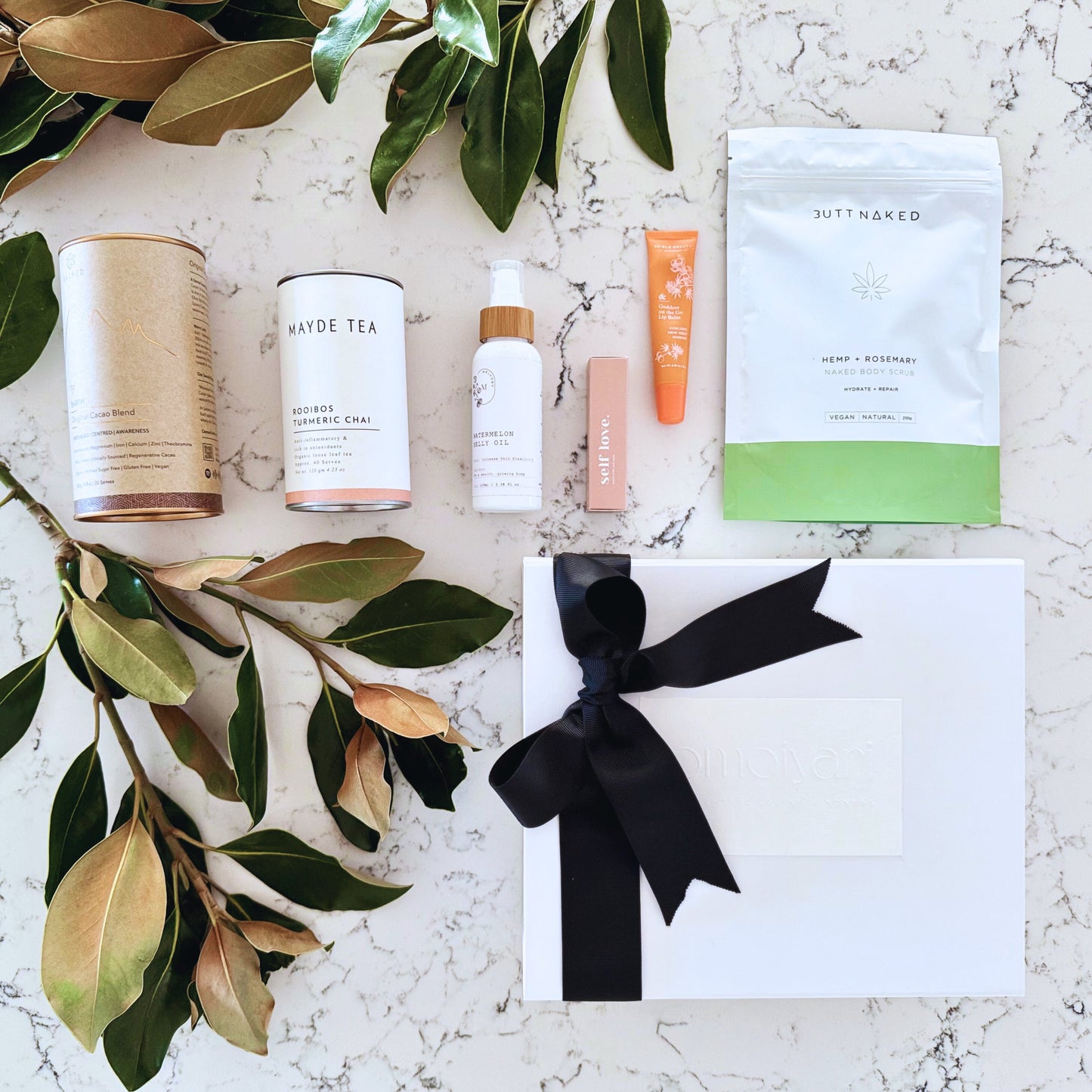 Omoiyari Glow & Grow gift hamper box - Skincare products, organic cacao and tea, stretch mark oil, lip balm and green magnolia leaves on a marble surface