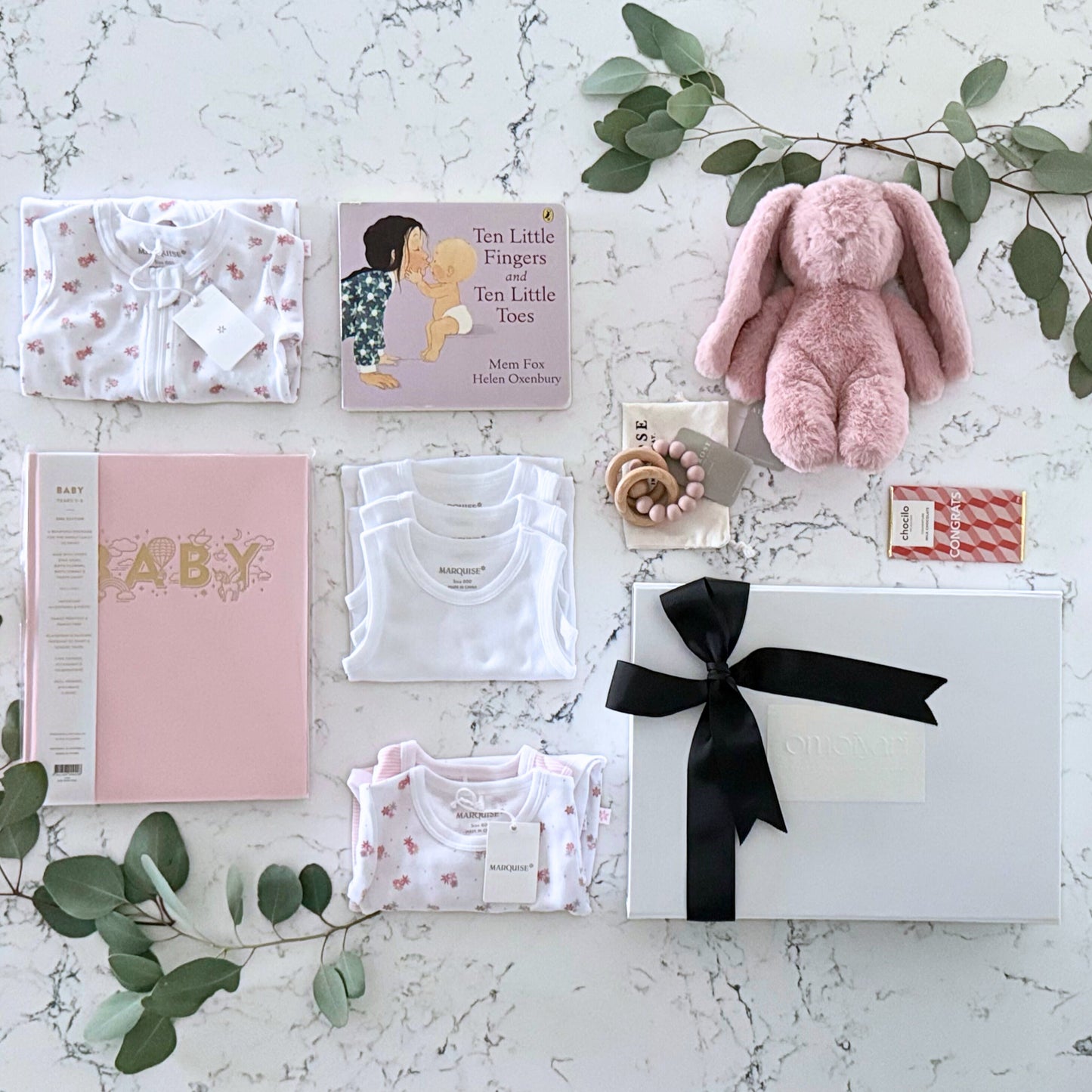 Omoiyari Hello Baby Girl Deluxe Gift hamper bod - Baby gift set with pink plush toy, Marquise baby clothes, teething ring on a marble surface with green leaves.