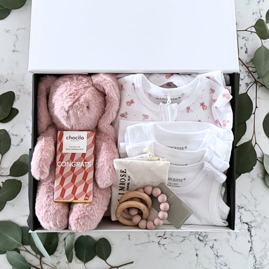 Omoiyari Hello Baby Girl Deluxe Gift hamper bod - Baby gift set with pink plush toy, Marquise baby clothes, teething ring on a marble surface with green leaves.