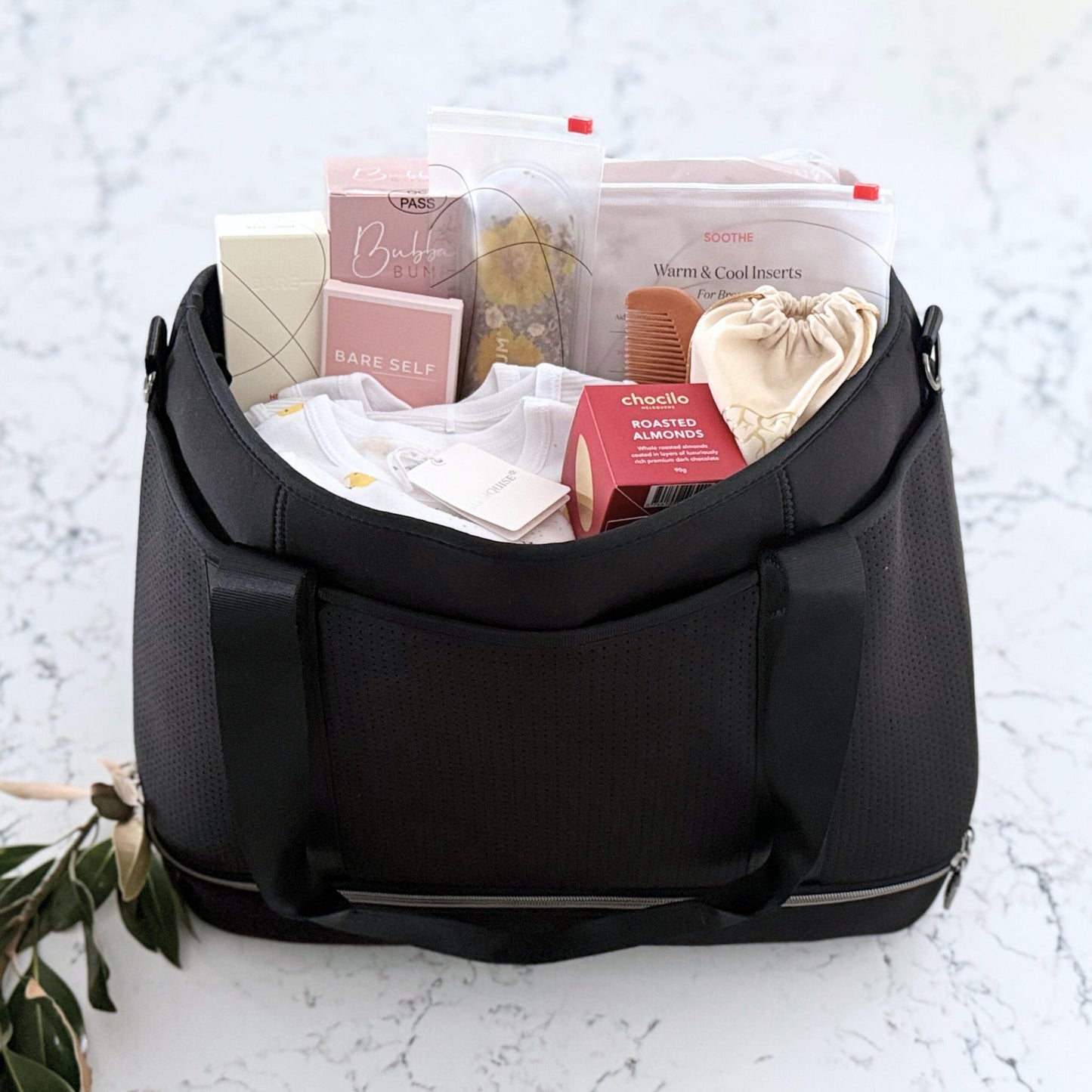 Omoiyari Hospital Bag Luxe with Prene Saturday Bag filled with Marquise babywear, bamboo nappies and wipes, perineal bottle, nipple cream, Essentials Oils roller, Postpartum disposable underwear, bra cooling inserts and chocolate on a marble surface with flowers.