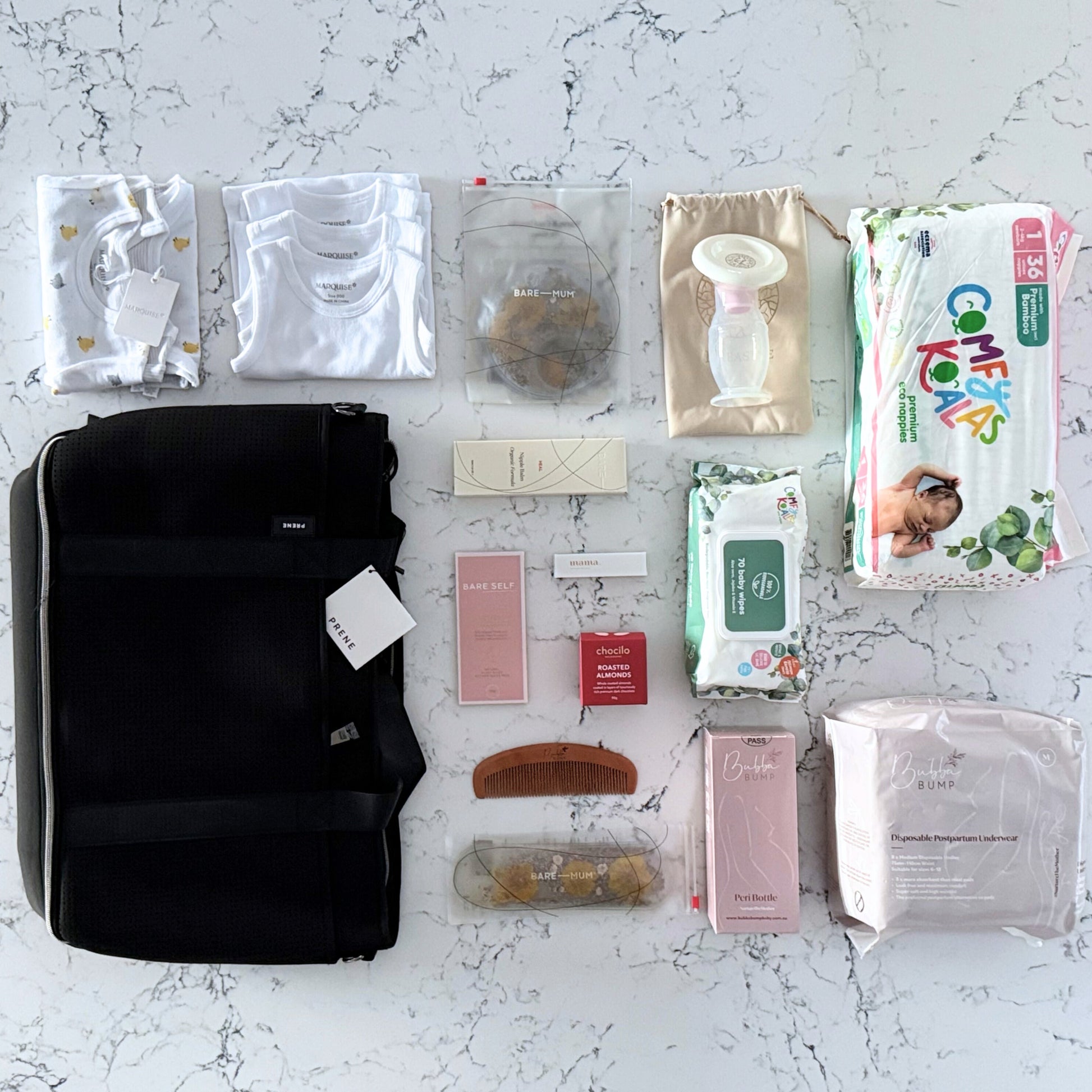 Omoiyari Hospital Bag Luxe with Prene Saturday Bag filled with Marquise babywear, bamboo nappies and wipes, perineal bottle, nipple cream, Essentials Oils roller, Postpartum disposable underwear, bra cooling inserts and chocolate on a marble surface with flowers.