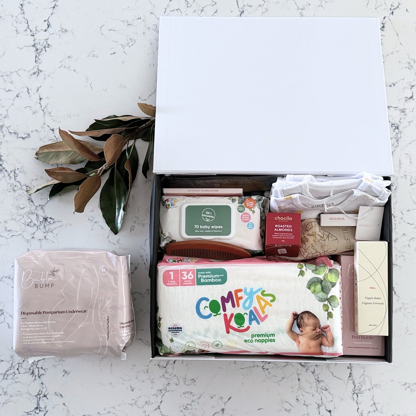 Omoiyari Hospital Bag Luxe gift hamper box with Marquise babywear, bamboo nappies and wipes, perineal bottle, nipple cream, Essentials Oils roller, Postpartum disposable underwear, bra cooling inserts and chocolate on a marble surface with flowers.