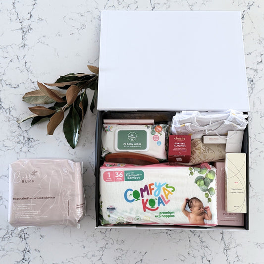 Omoiyari Hospital Bag Luxe gift hamper box with Marquise babywear, bamboo nappies and wipes, perineal bottle, nipple cream, Essentials Oils roller, Postpartum disposable underwear, bra cooling inserts and chocolate on a marble surface with flowers.