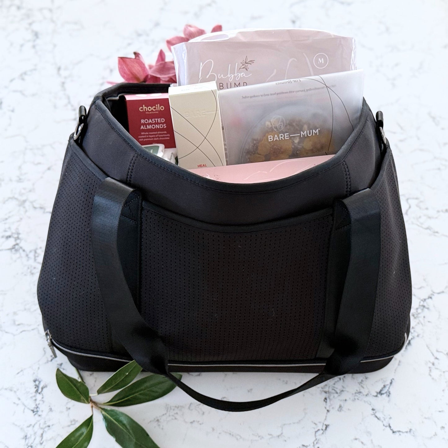 Omoiyari Hospital Bag with Prene Saturday Bag with bamboo nappies and wipes, perineal bottle, nipple cream, Essentials Oils roller, Postpartum disposable underwear, bra cooling inserts and chocolate on a marble surface with flowers.