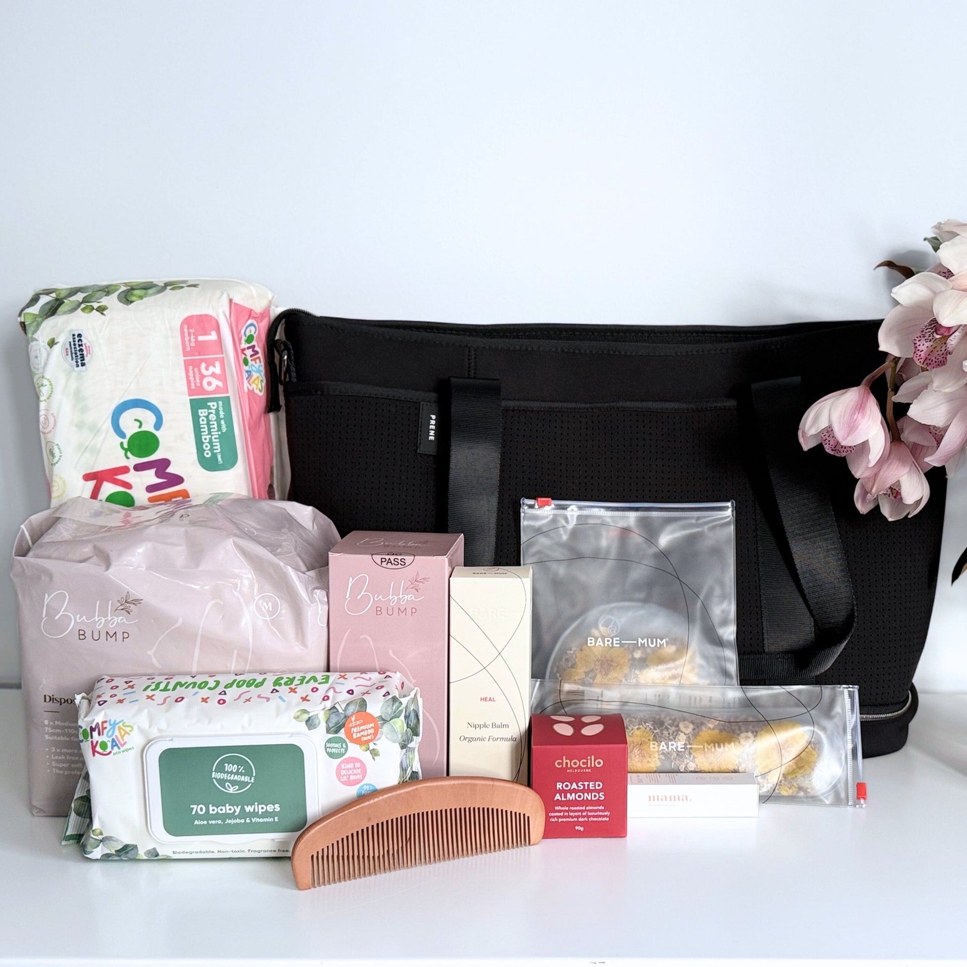 Omoiyari Hospital Bag with Prene Saturday Bag with bamboo nappies and wipes, perineal bottle, nipple cream, Essentials Oils roller, Postpartum disposable underwear, bra cooling inserts and chocolate on a marble surface with flowers.