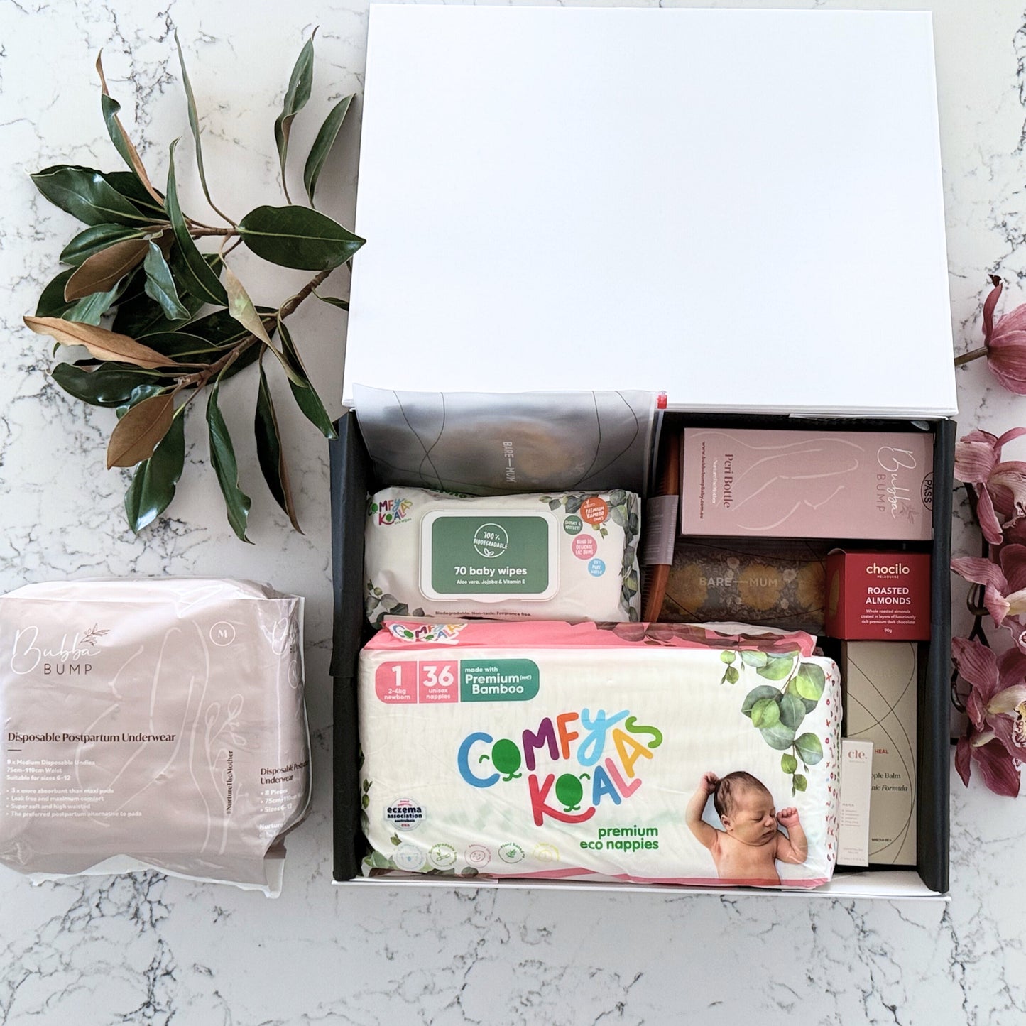 Omoiyari Hospital Bag gift hamper box  with bamboo nappies and wipes, perineal bottle, nipple cream, Essentials Oils roller, Postpartum disposable underwear, bra cooling inserts and chocolate on a marble surface with flowers.